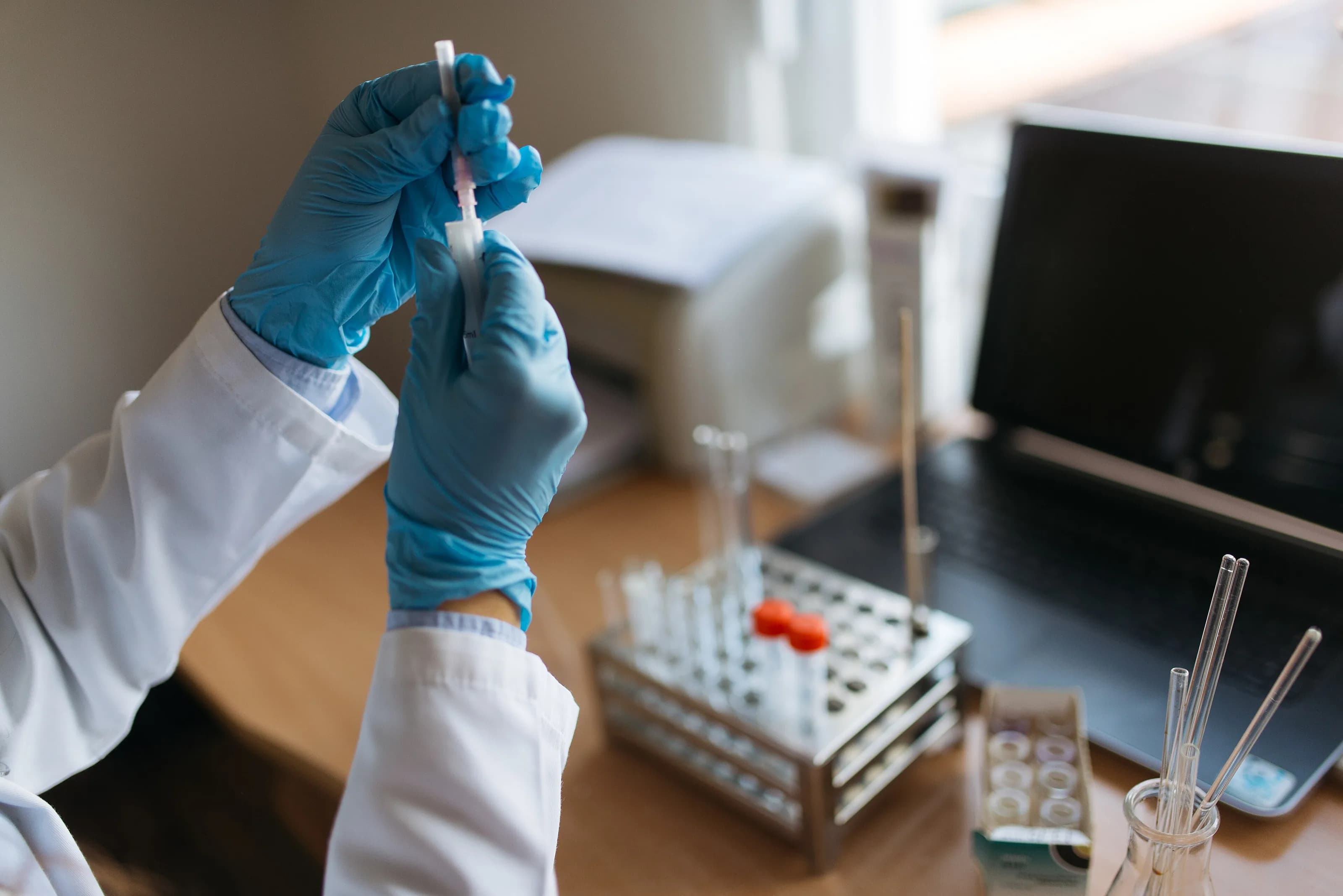 Manos con guantes y una jeringa junto a tubos de ensayo en un laboratorio